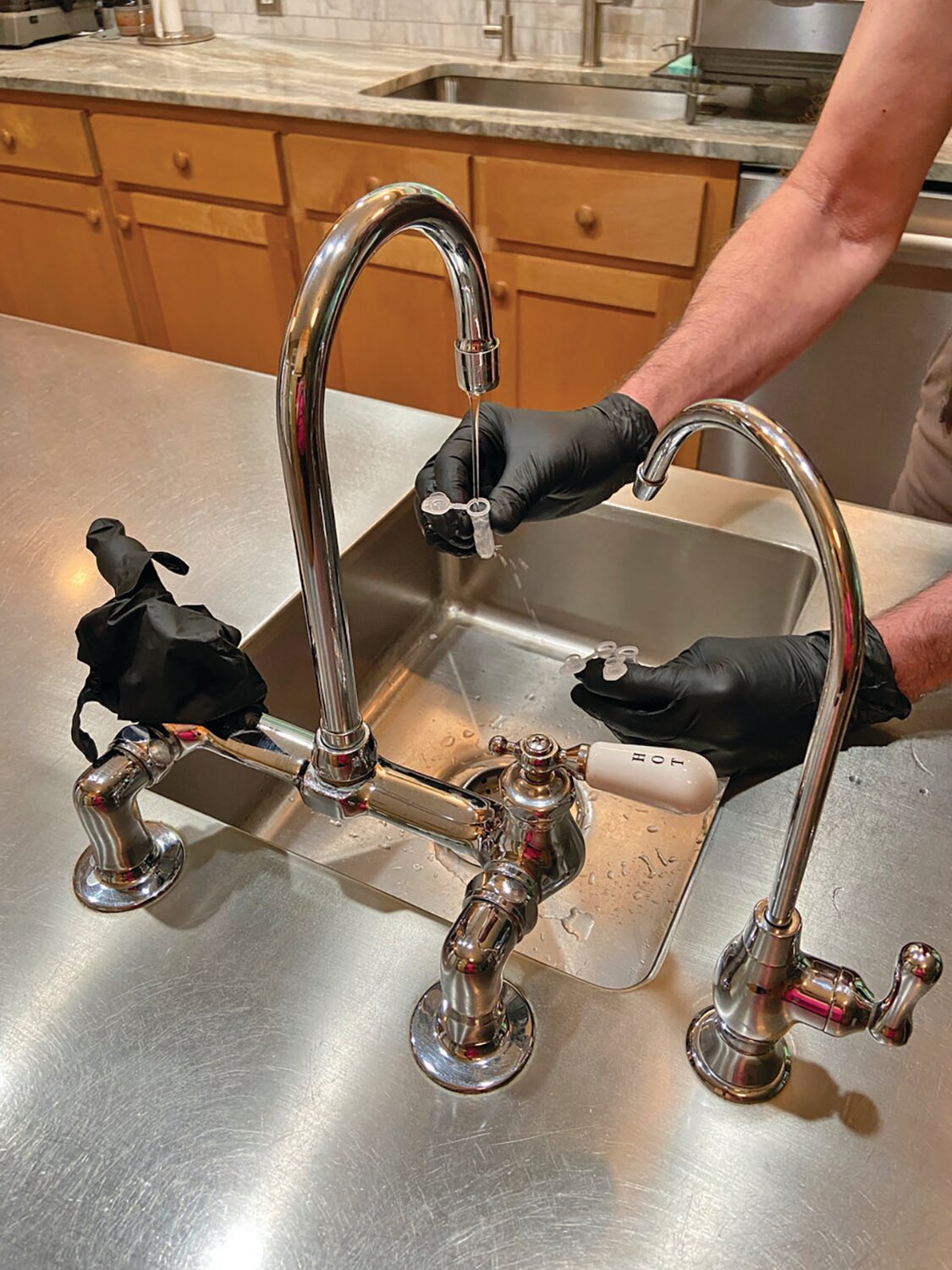 A USGS scientist wearing black gloves is collecting a sample of tap water from the kitchen sink using small plastic vials to test for PFAS.